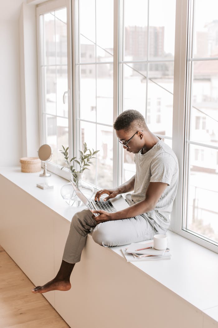 A man sits on a window sill, using a laptop for remote work or study, surrounded by minimal decor.
