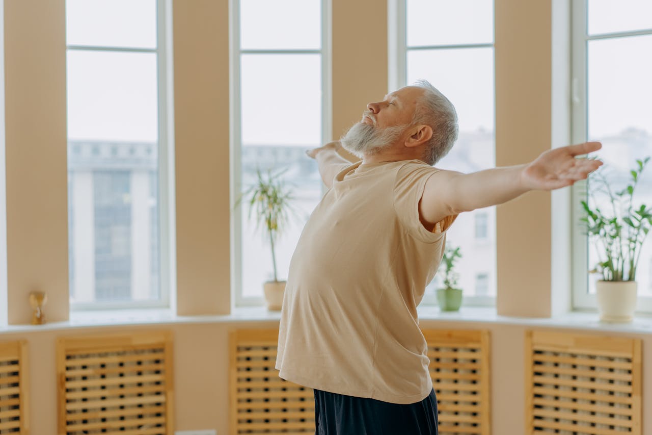 Elderly man meditating with eyes closed, arms wide in an indoor space, expressing calm.